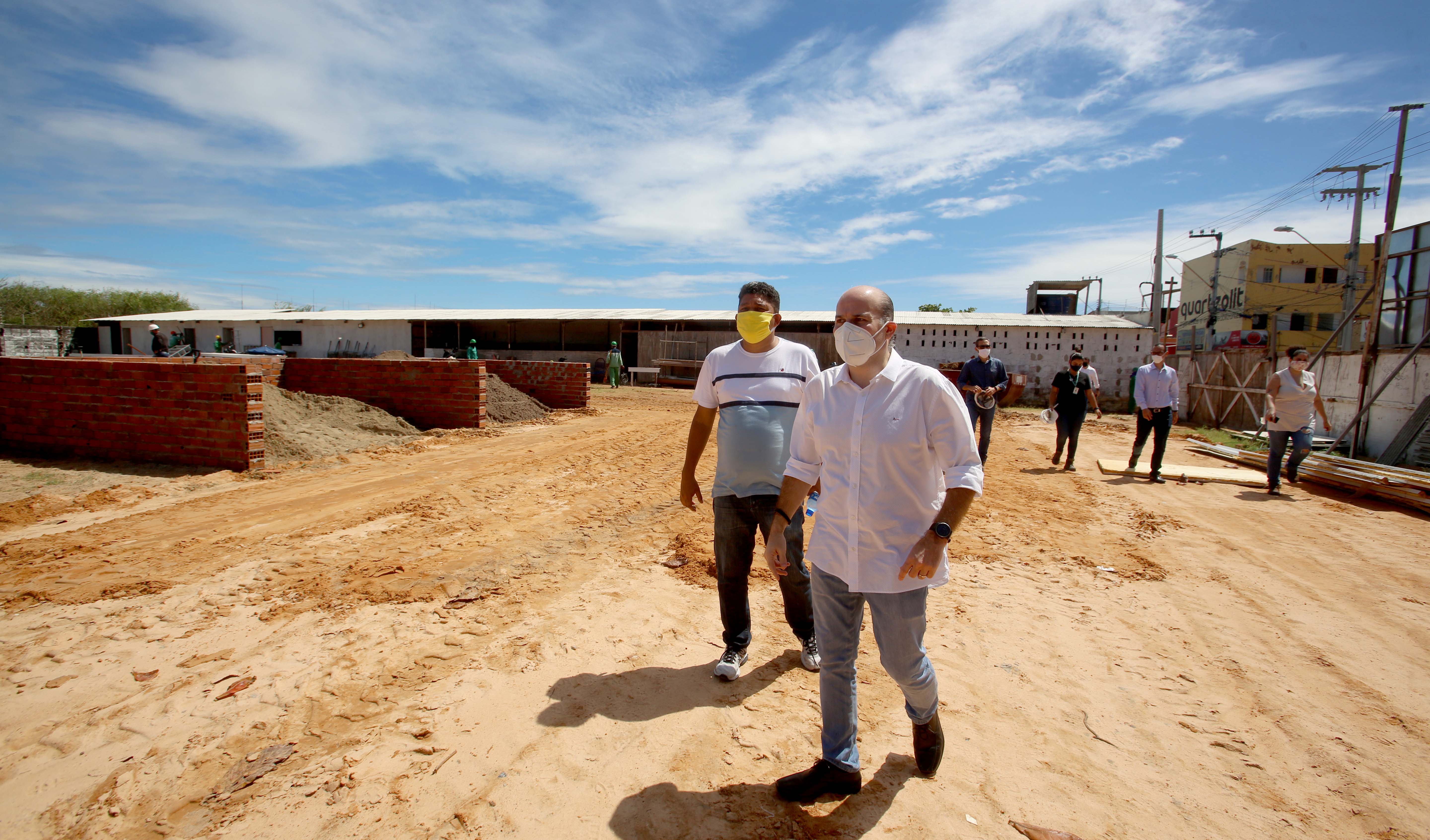 prefeito de máscara caminhando por terreno com obra ao fundo e homem de máscara andando ao seu lado